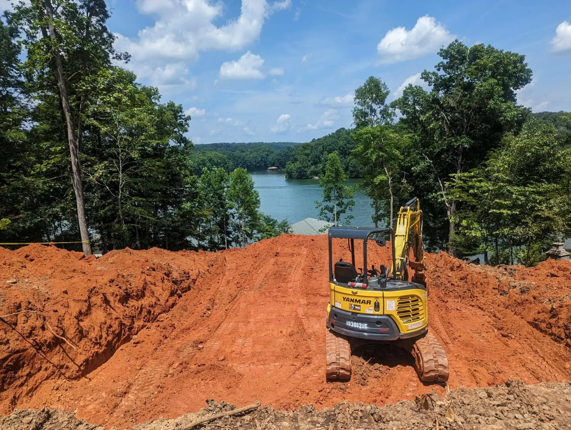 Mini excavator cutting a building pad in red clay overlooking a lake
